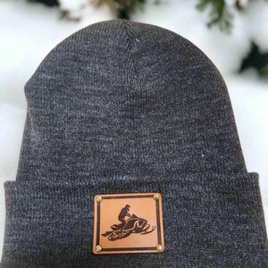 May include: A dark gray beanie hat with a brown leather patch featuring a snowmobile design. The patch is secured with four brass rivets. The hat is made of a knitted material.
