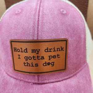 May include: A pink trucker hat with a white mesh back. A brown leather patch on the front displays the text "Hold my drink I gotta pet this dog" in black lettering. The hat has a curved brim and a button on top.