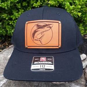 May include: Black trucker hat with a leather patch featuring a shark illustration. The patch is secured with brass rivets. The hat includes a mesh back and a curved brim. The Richardson brand label is visible.