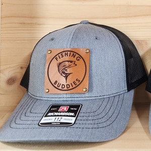 May include: A gray trucker hat with a black mesh back. The front features a brown leather patch with the words "FISHING BUDDIES" around a fish graphic. The hat has a curved brim and a "RICHARDSON" tag.