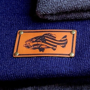Pode incluir: Uma touca azul marinho com um patch de couro castanho. O patch apresenta um desenho de peixe preto com listras da bandeira americana, fixado com rebites de latão. Uma touca cinza dobrada é visível no fundo.