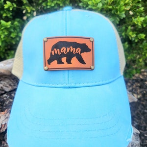 Pode incluir: Boné de caminhoneiro azul claro com aba desgastada. O boné apresenta uma parte traseira de malha bege e um patch de couro marrom com a palavra "mama" e uma silhueta de urso.