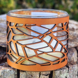 May include: A candle holder with a clear glass jar and a decorative, openwork, orange-brown leather sleeve. The sleeve features a geometric pattern of interconnected shapes. The candle inside is white, and the holder sits on a textured, brown surface.