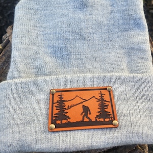 May include: A pale grey beanie hat with a brown leather patch. The patch depicts a mountain scene with trees and a Bigfoot silhouette. The patch is secured with four brass rivets. The hat is made of a soft, knitted material.