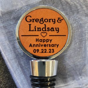 May include: A silver-toned bottle stopper featuring a circular orange insert with black engraved text that reads Gregory & Lindsay, a heart symbol, Happy Anniversary, and the date 09.22.23, finished with a black ribbed rubber base.