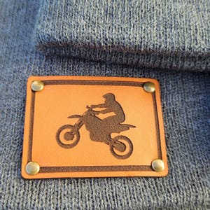 May include: A blue knit beanie with a rectangular, tan leather patch. The patch features a dark brown silhouette of a person riding a dirt bike, secured with four brass rivets. The beanie is a solid color.