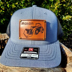 Pode incluir: Boné trucker azul com parte traseira em rede preta. Um patch de couro castanho exibe o nome "Mason" acima de uma ilustração de uma carregadeira frontal. O boné inclui uma etiqueta "Richardson".