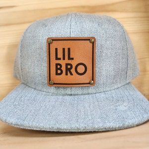 Puede incluir: Gorra de béisbol gris claro con visera plana. Un parche cuadrado de cuero marrón está centrado en la parte delantera, asegurado por cuatro remaches de latón. El parche presenta las palabras "LIL BRO" en negrita, fuente sans-serif.