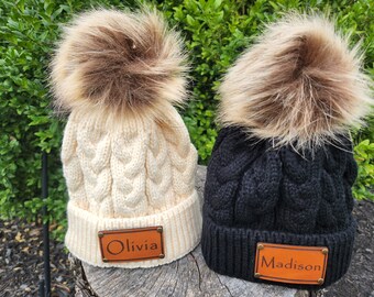 Peut inclure: Deux bonnets en tricot torsadé avec des pompons en fausse fourrure. L'un est crème, l'autre noir. Chacun a un patch en similicuir marron avec un nom : "Olivia" et "Madison". Les chapeaux sont exposés sur une surface en bois.