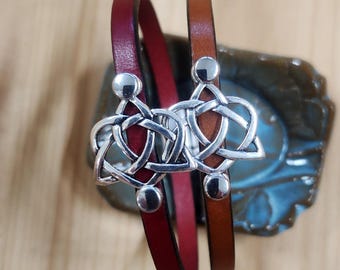Op de afbeelding: Twee leren armbanden, één bordeauxrood en één bruin, zijn versierd met zilveren Keltische knoopbedels. Elke armband heeft een zilveren bedel met een Keltisch knoopontwerp en zilveren balaccenten. De armbanden worden gepresenteerd op een getextureerd oppervlak.
