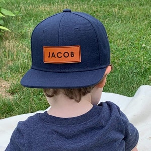Puede incluir: Gorra de béisbol azul marino con visera plana. La gorra tiene un parche rectangular de cuero marrón con el nombre "JACOB" en letras mayúsculas negras. La gorra la lleva una persona con una camiseta azul marino.