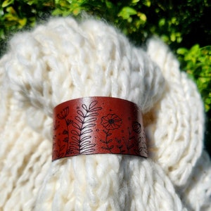 May include: A brown leather cuff bracelet with a floral design. The bracelet has black line art of flowers and foliage. The cuff is displayed on a white, chunky knit fabric, with green foliage in the background.