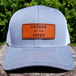 Peut inclure: Casquette de camionneur grise avec un dos en filet blanc et un patch en cuir marron. Le patch porte l'inscription "FATHER OF THE BRIDE" en noir. La casquette est posée sur une surface en bois.