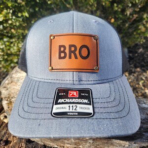 Puede incluir: Gorra de camionero gris con parte trasera de malla negra. Cuenta con un parche de cuero marrón con la palabra "BRO" en relieve. La gorra tiene una visera curva y una etiqueta Richardson Original 112 Youth.