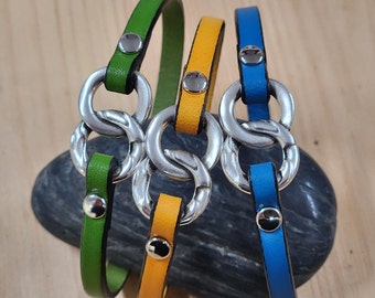 Op de afbeelding: Leren armbanden in groen, geel en blauw. Elke armband heeft een zilverkleurig, in elkaar grijpend ringontwerp en een drukknoopsluiting. De armbanden worden gepresenteerd op een houten oppervlak en een donkergrijze steen.