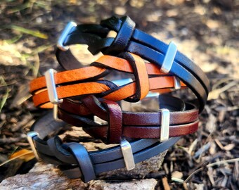 Op de afbeelding: Vier leren armbanden in zwart, oranje, bordeauxrood en grijs. Elke armband heeft een gevlochten ontwerp met een zilverkleurige metalen sluiting. De armbanden zijn op elkaar gestapeld en tonen hun verschillende kleuren en texturen.