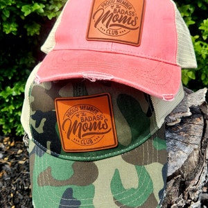 Pode incluir: Dois bonés de beisebol. Um é rosa com as costas em malha bege, o outro é camuflado com as costas em malha bege. Ambos têm um patch de couro com o texto "PROUD MEMBER of the BADASS MOMS CLUB".