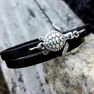 Könnte beinhalten: Ein schwarzes Lederarmband mit einem silberfarbenen Schildkrötenanhänger. Der Schildkrötenanhänger ist mit strukturierten Schuppen versehen und hat einen Hakenverschluss. Das Armband wird zweimal um das Handgelenk gewickelt.