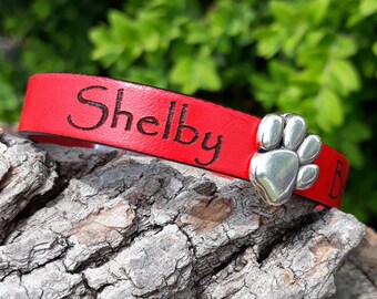 May include: A red leather dog collar with the name "Shelby" engraved. A silver paw print charm is attached. The collar rests on textured wood. The background is blurred green foliage.