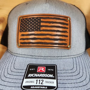 Op de afbeelding: Grijze truckerpet met een zwarte mesh achterkant en een leren patch met een Amerikaanse vlag. De patch is bevestigd met messing klinknagels. De pet heeft een gebogen klep en een verstelbare band.
