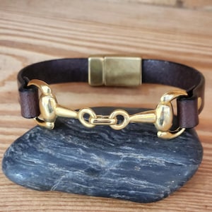 Puede incluir: Una pulsera de cuero marrón con un diseño de bocado de caballo dorado. La pulsera tiene un cierre magnético y se muestra sobre una piedra gris oscura. La banda de cuero mide aproximadamente 2 cm de ancho.