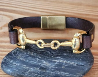 May include: A dark brown leather bracelet featuring a polished gold-tone metal horse bit centerpiece and a rectangular magnetic clasp. The leather band is secured to the metal bit on both sides, creating a continuous loop design for the wrist.