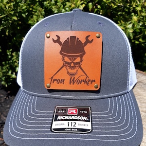 May include: A gray trucker hat with a white mesh back and a leather patch. The patch features a skull wearing a hard hat, crossed wrenches, and the words "Iron Worker". The hat has a curved brim and white stitching.