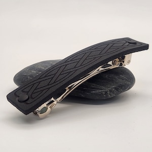 May include: A black leather barrette with a Celtic knot design. The barrette features a silver-colored metal clasp and a button closure. The hair accessory is approximately 4 inches long.