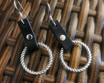 May include: A pair of black leather earrings with silver-colored twisted hoops. The earrings have a rectangular black leather top with a silver button. The hoops are a twisted rope design and hang from silver ear wires.
