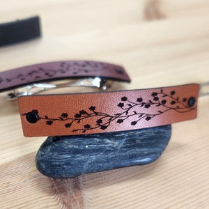 Puede incluir: Dos horquillas de cuero. Una es marrón, la otra es marrón rojizo. Cada una tiene un diseño floral negro y un broche negro. Las horquillas están sobre una superficie de madera.