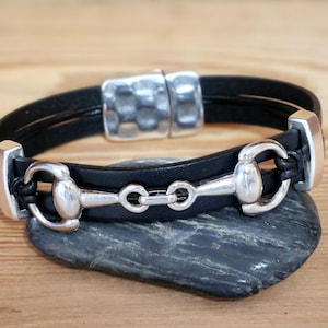 May include: Black leather bracelet with silver-tone equestrian bit and buckle. The bracelet is displayed on a dark gray stone against a wooden background. The bracelet features a magnetic clasp.
