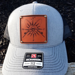 Op de afbeelding: Een grijze truckerpet met een zwarte mesh achterkant en een bruine leren patch. De patch heeft een kompasontwerp met een schedel en hengels. De pet heeft de tekst "RICHARDSON" en "ORIGINAL 112 TRUCKER".