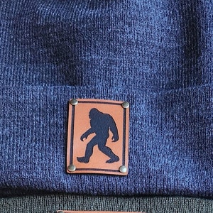 Puede incluir: Un gorro de punto azul marino con un parche de cuero marrón que presenta una silueta negra de Bigfoot. El parche está asegurado con cuatro pequeños remaches metálicos. El gorro está doblado en el borde.