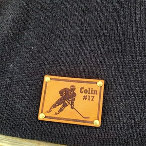 May include: A dark navy beanie with a rectangular, tan leather patch. The patch features a hockey player graphic, the name "Colin," and the number "17." The patch is secured with four brass rivets.