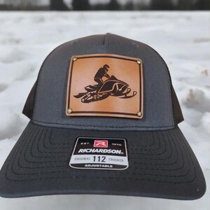 May include: A gray Richardson 112 trucker hat with a brown leather patch featuring a snowmobiler graphic. The hat has a curved brim and adjustable closure. The patch is secured with brass rivets.
