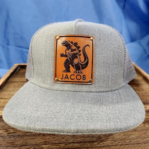 May include: Gray trucker hat with a brown leather patch featuring a Godzilla graphic. The patch is secured with brass rivets and includes the name "JACOB" in white lettering. The hat has a curved brim and mesh back.