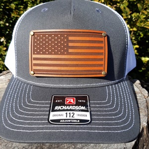 Op de afbeelding: Een grijze en witte truckerpet met een bruinleren Amerikaanse vlag patch. De pet heeft een gebogen klep en een mesh achterkant. De patch is bevestigd met messing klinknagels. De Richardson Original 112 Trucker pet is verstelbaar.