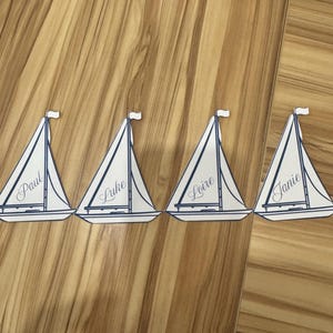 Könnte beinhalten: Vier Platzkarten in Segelbootform mit in Kursivschrift geschriebenen Namen: Paul, Luke, Loire und Janie. Jede Karte hat ein weißes Segel mit blauem Rand und eine kleine weiße Flagge oben. Die Karten sind auf einer Holzoberfläche angeordnet.