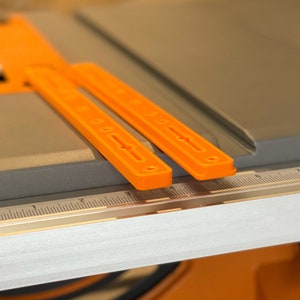 May include: Two orange plastic rulers with markings on a gray surface. The rulers are aligned with a silver measuring tape.