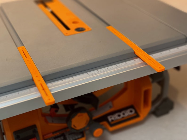 May include: A gray table saw with orange plastic rulers and a black and orange base. The table saw has a ruler with markings in inches.