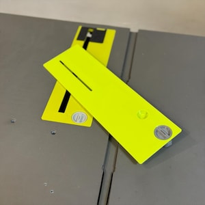May include: Two bright yellow table saw accessories are shown. One is positioned on top of the other. Each has a slot and a circular detail. The accessories are on a gray surface.