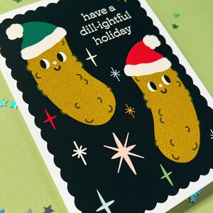 Pu&ograve; includere: Un biglietto nero con un bordo bianco a festoni. Il biglietto ha uno sfondo nero con testo bianco che dice "have a dill-ightful holiday". Ci sono due ornamenti di cetriolo verdi e rossi con delle facce. Gli ornamenti indossano cappelli di Babbo Natale.