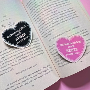 May include: Two heart-shaped stickers with text. The first sticker is black with white text that reads "my book boyfriend would NEVER do this to me". The second sticker is pink with white text that reads "my book boyfriend would NEVER do this to me".