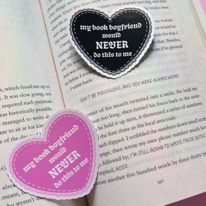 May include: Two heart-shaped stickers with the text "my book boyfriend would NEVER do this to me". One sticker is pink with black text and the other is black with white text. The stickers are placed on a book page with text.