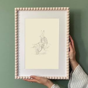 May include: A framed black and white illustration of a rabbit pulling a wheelbarrow. The frame is light pink with a beaded border.