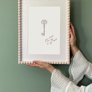 Puede incluir: Un marco blanco con un borde de cuentas rodea una impresión de una llave plateada con el texto "you hold the key to my heart" escrito en tinta rosa.