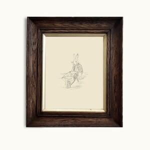 May include: A framed black and white pencil drawing of a rabbit pushing a wheelbarrow. The drawing is on a cream-colored paper and is framed in a dark wood frame.