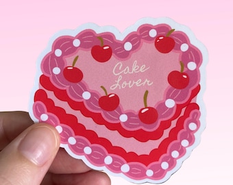 May include: A pink heart-shaped cake with white frosting and red cherries. The cake has the words "Cake Lover" written on it.