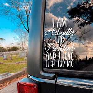 Könnte beinhalten: Ein weißer Vinyl-Aufkleber auf einer Fahrzeugscheibe mit dem Text "I AM Actually THE Problem AND I LOVE THAT FOR ME" in einer Kombination aus Schreibschrift und serifenloser Schrift, verziert mit kleinen vierzackigen Sternsymbolen.