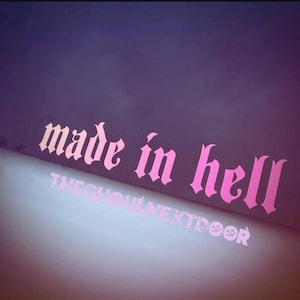 Peut inclure: Texte rose sur fond violet dégradé : "made in hell" en police gothique. En dessous, le texte plus petit dit "THEGHOULNEXTDOOR" avec une citrouille souriante et un crâne.